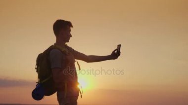 Bir genç bir sırt çantası ile kendini bir telefon ile resimler. Batan güneşin karşı. Turizm ve seyahat