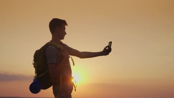 Un adolescente con una mochila se imagina a sí mismo con un teléfono. Contra el sol poniente. Turismo y viajes — Vídeo de stock