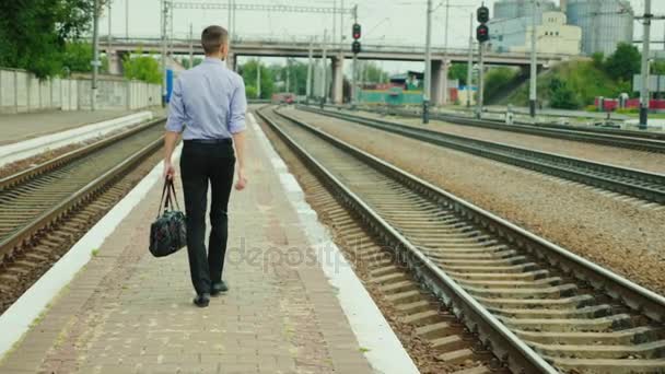 Rear view: A young successful businessman walks along the railway station along the tracks. He carries a bag in his hand, smiles. A successful business trip — Stock Video