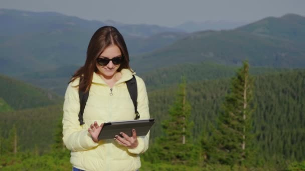 Una turista con una chaqueta amarilla usa una tableta. Se alza sobre el fondo de las montañas cubiertas de bosque. Forma activa de vida y tecnología — Vídeo de stock