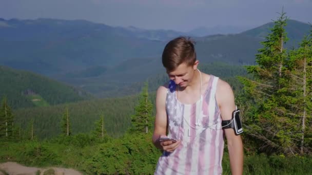 Joven atleta masculino usando el teléfono. Se encuentra sobre un hermoso telón de fondo de montañas y bosques. Aplicación deportiva para smartphone — Vídeo de stock