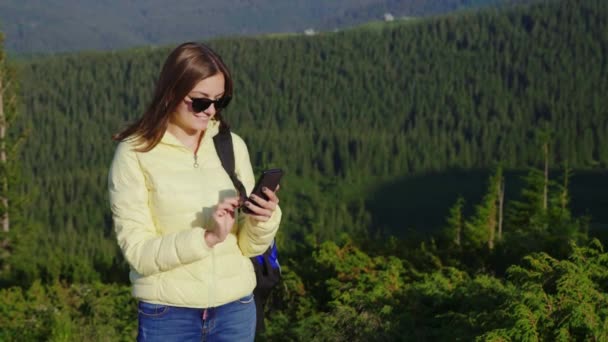 Mujer joven disfrutando de un smartphone en un pintoresco telón de fondo de montañas cubiertas de bosque. Siempre en línea — Vídeos de Stock