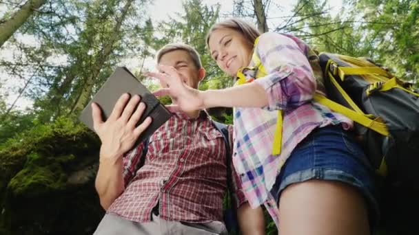 Una giovane coppia di turisti con gli zaini si godono un tablet durante un viaggio. Stanno nella foresta, il sole splende splendidamente dall'alto. Navigazione e ricerca direzione — Video Stock