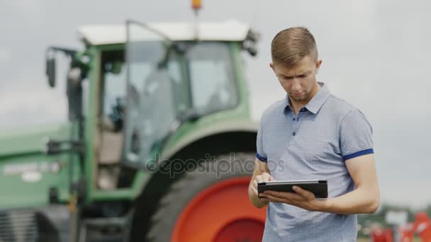 Egy fiatal kaukázusi mezőgazdasági termelő dolgozik egy tabletta területén. A háttérben egy nagy traktor — Stock videók