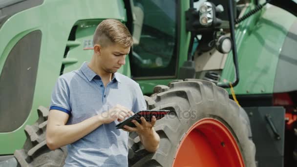 Un granjero exitoso está trabajando en el campo. Utiliza una tableta, se para cerca del tractor — Vídeos de Stock