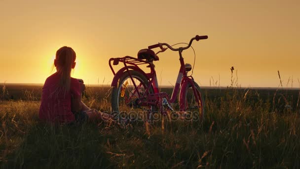 A menina tem 6 anos sentada perto da bicicleta. Usa um smartphone. Local pitoresco ao pôr-do-sol — Vídeo de Stock