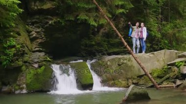 Turist genç bir çift bir dağ nehir üzerinde bir şelale yakınındaki bekliyoruz. Güzel sahne hayran. Turizm ve aktif yaşam tarzı