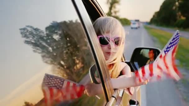 Una niña de 6 años con una bandera americana mira por la ventana de un coche en movimiento — Vídeo de stock