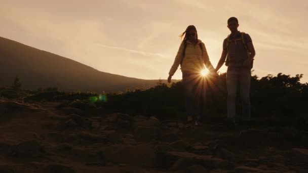 Egy fiatal pár, a turisták, hátizsákok, gyaloglás a hegyi ösvényen. A lenyugvó nap sugaraiban — Stock videók