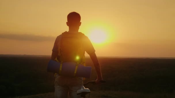 Un hombre con una mochila y una bicicleta. Se para y mira al horizonte donde se pone el sol. Ciclismo y aventura — Vídeos de Stock