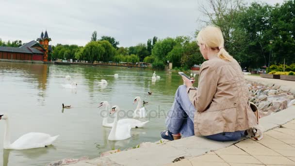 Una giovane donna siede sul lago con dei cigni. Utilizza uno smartphone . — Video Stock