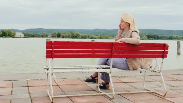 Μια γυναίκα μιλάει στο τηλέφωνο. Κάθεται σε ένα κόκκινο παγκάκι στο φόντο της το γραφικό λίμνη Μπάλατον στην Ουγγαρία — Αρχείο Βίντεο