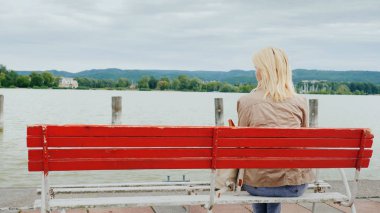 Bir kadının göle manzaranın kırmızı bir bankta oturur. Arkadan Görünüm. Lake Balaton Macaristan üzerine getirin