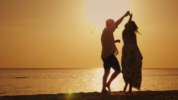 Силуэты молодой пары, забавные танцы на фоне моря и заходящего солнца. Веселый праздник у моря — стоковое видео