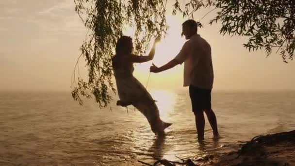 Siluetele unui cuplu tânăr, un bărbat își rostogolește soția pe un leagăn. Pe fundalul mării la apusul soarelui. Plouă — Videoclip de stoc