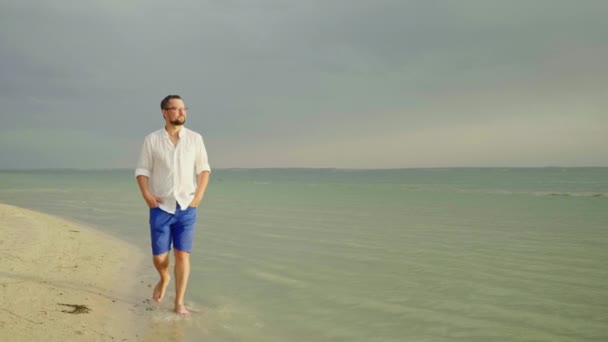 Młodych, dobrze wyglądający mężczyzna w lekkie ubrania i okulary. Idzie się wzdłuż plaży nad brzegiem morza. Uciec od trosk, wolność i urlopu. 4 k film w zwolnionym tempie — Wideo stockowe
