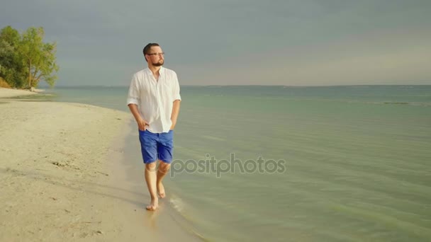 Un homme solitaire en vêtements légers se promène pieds nus le long du sable le long de la mer. Fuyez les soucis, la liberté et partez. Vidéo au ralenti 4K — Video