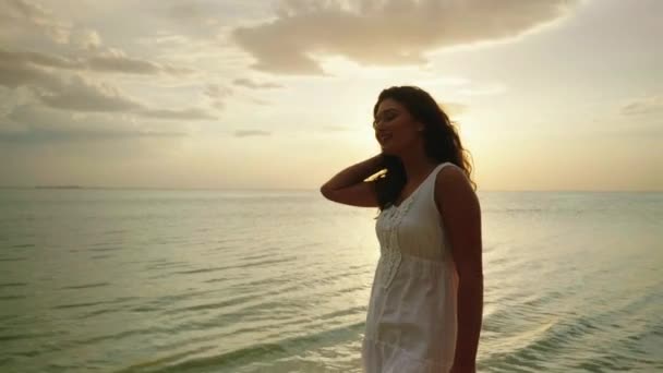 Молодая женщина в светлом летнем платье идет по краю воды на фоне моря и заката. Отличный отпуск. 4K замедленное видео — стоковое видео