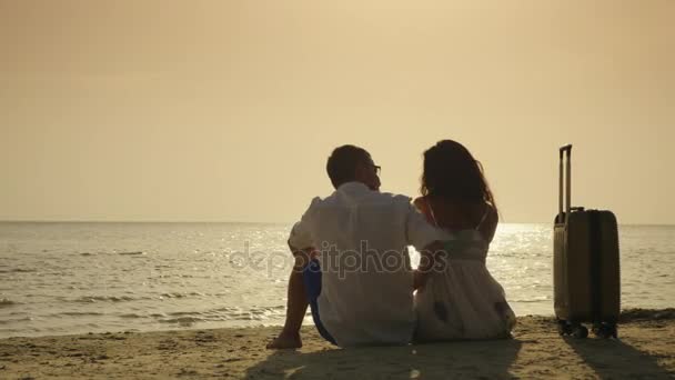 休暇に到着。若いカップルは旅行バッグの近くの砂の上に座っています。海に夕日を楽しんでください。 — ストック動画