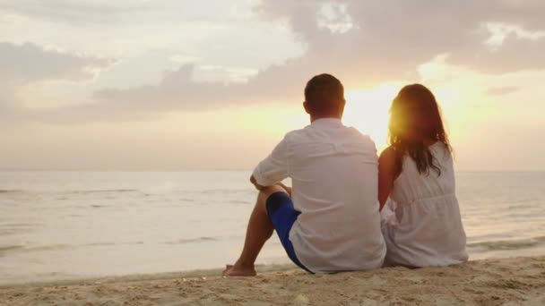 Egy férfi és egy nő egymás mellett ül a homok a tengerparton. Együtt néznek a napnyugtakor a tenger felett — Stock videók