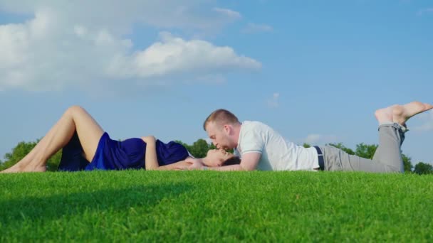 Mladý muž jemně políbí svou těhotnou manželku. Společně leží na zeleném trávníku v parku proti modré obloze — Stock video