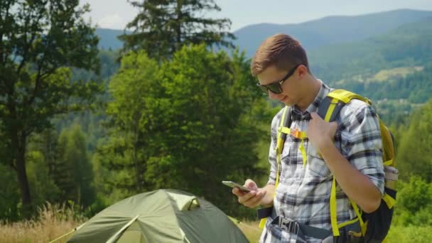 Ein junger Mann mit Rucksack benutzt auf dem Campingplatz ein Smartphone. immer in Kontakt, Anwendungen für den Tourismus — Stockvideo