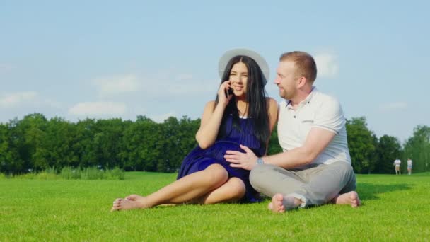 Młoda kobieta w ciąży mówi przez telefon. Obok niej siedzi jej mąż, reszta razem w parku — Wideo stockowe