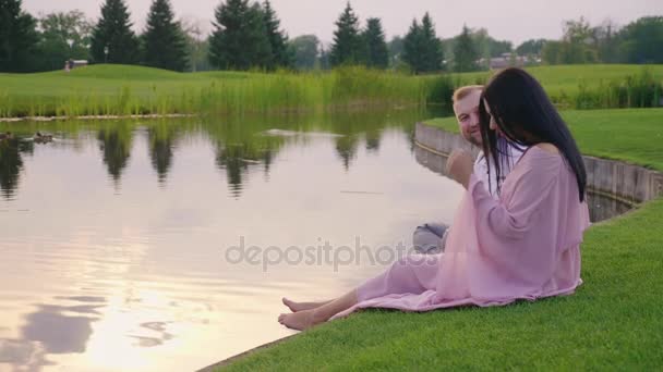 Egy terhes nő, egy rózsaszín ruhás férjével. Ők ül a fűben, a tó partján — Stock videók