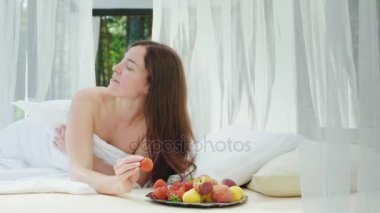 Bir çilek onun elinde olan genç kadın yatakta. Bir tembel olarak havuzun perdeler ile yatıyor. Romantik balayı, yatakta kahvaltı