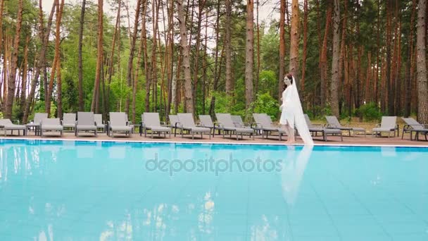La sposa con un lungo velo sta camminando intorno alla piscina. Matrimonio in un hotel di lusso — Video Stock
