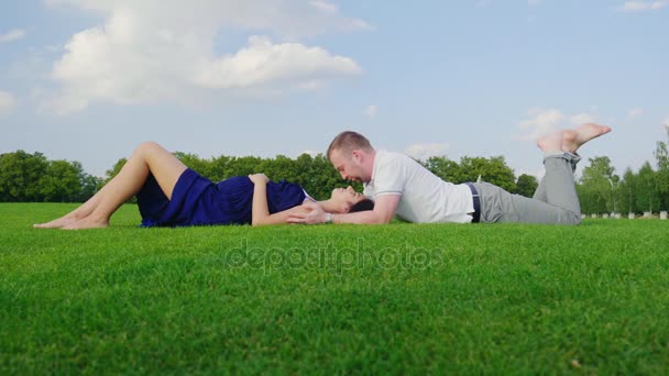 若い家族は、子供を待っています。男が妊娠中の妻にキス、一緒に公園で芝生に寝転んで — ストック動画
