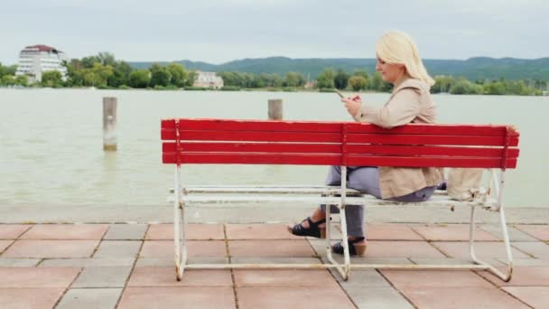 La mujer se sienta en un banco en el fondo de un gran lago. Usa un teléfono móvil. Lago Balaton en Hungría — Vídeos de Stock