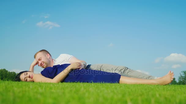 Молодой муж с беременной женой отдыхают в парке, вместе лежат на зеленой траве — стоковое видео