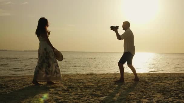 En strand-fotograf tar bilder på en ung kvinna mot bakgrund av havet och solnedgång. — Stockvideo