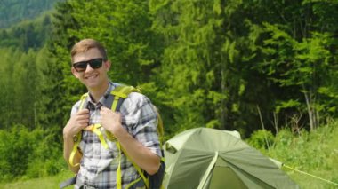 Genç bir adam bir sırt çantası ile bir kamp çadır arka planda duran kameraya bakıyor. Bir gezgin portresi