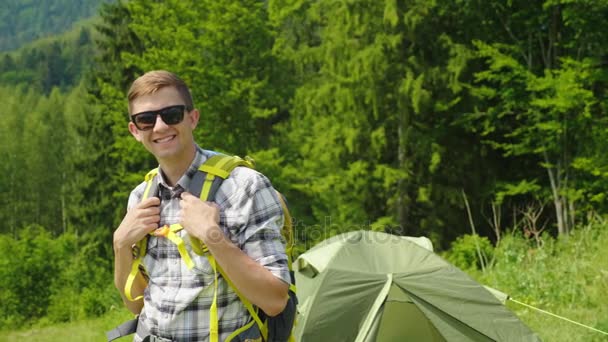Um jovem com uma mochila está olhando para a câmera, de pé no fundo de uma tenda de acampamento. Retrato de um viajante — Vídeo de Stock