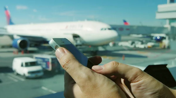 Manos de un hombre con un smartphone. Está escribiendo texto, utiliza aplicaciones. En el fondo de una ventana en el aeropuerto donde se pueden ver los aviones —  Fotos de Stock