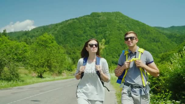 Un jeune couple de touristes se promène le long de la route vers de belles montagnes couvertes de forêt. Mode de vie actif et vacances — Video
