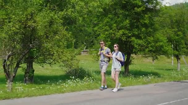 Seitenansicht: Ein junges Touristenpaar geht die Straße entlang zu wunderschönen, bewaldeten Bergen. Aktive Lebensweise und Urlaub — Stockvideo