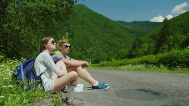 Un par de turistas cansados con mochilas descansan en la carretera. En un lugar pintoresco en el fondo de las montañas . — Vídeo de stock