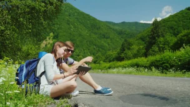 Un par de viajeros con mochilas se sienta al lado de la carretera. Descansan, usan una tableta digital — Vídeos de Stock