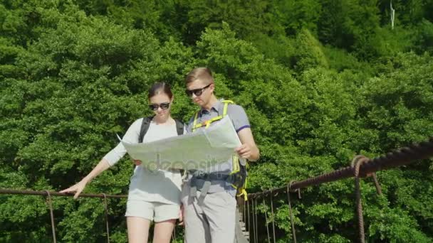 Un hombre y una mujer están estudiando el mapa. Se encuentran en el puente en el fondo de un pintoresco paisaje de verano . — Vídeos de Stock