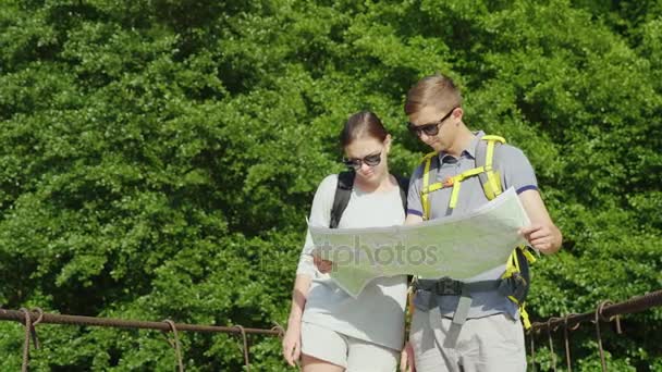 A man and a woman are studying the map. They stand on the bridge in the background of a picturesque summer landscape. — Stock Video