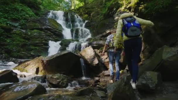 Un homme et une femme avec des sacs à dos escaladent les rochers jusqu'à une cascade — Video