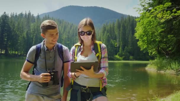 La pareja utiliza una tableta mientras viaja al lago y las montañas. Un hombre mira a través de los prismáticos, una mujer en un mapa en una tableta — Vídeos de Stock