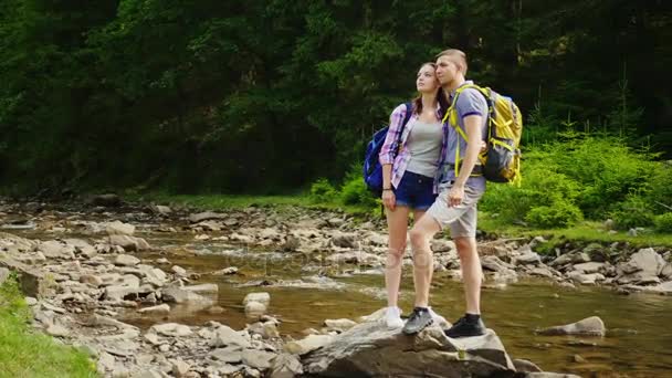 Влюбленная пара любуется прекрасным пейзажем, стоит на скале возле горной реки. Путешествие и активный образ жизни — стоковое видео