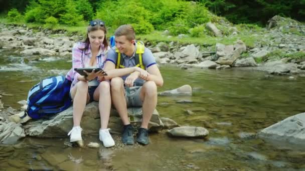 Mladý pár turistů si tablet na výletě. Sedí na skále poblíž horské řeky — Stock video