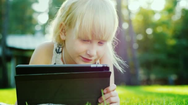 금발 소녀 6 년은 태블릿에 감정적으로 반응 한다. 집의 뒤뜰에 잔디에 누워 — 비디오