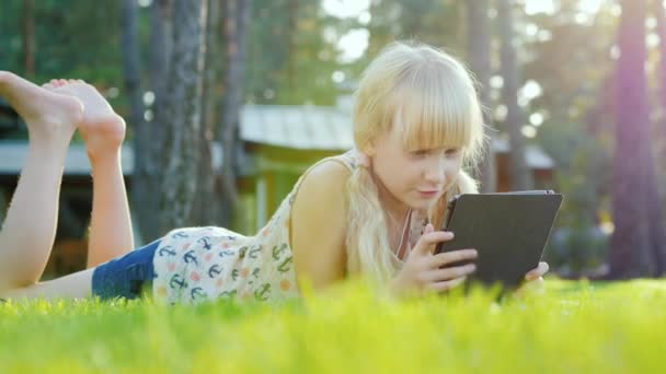 Blond meisje reageert 6 jaar spelen op de tablet, emotioneel. Liggend op het gazon in de achtertuin van het huis. Zijaanzicht — Stockvideo