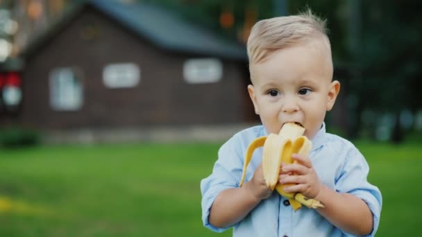 쾌활 한 1 년 된 아기는 바나나를 먹고 있다. 집의 뒤에 그것의 서 — 비디오